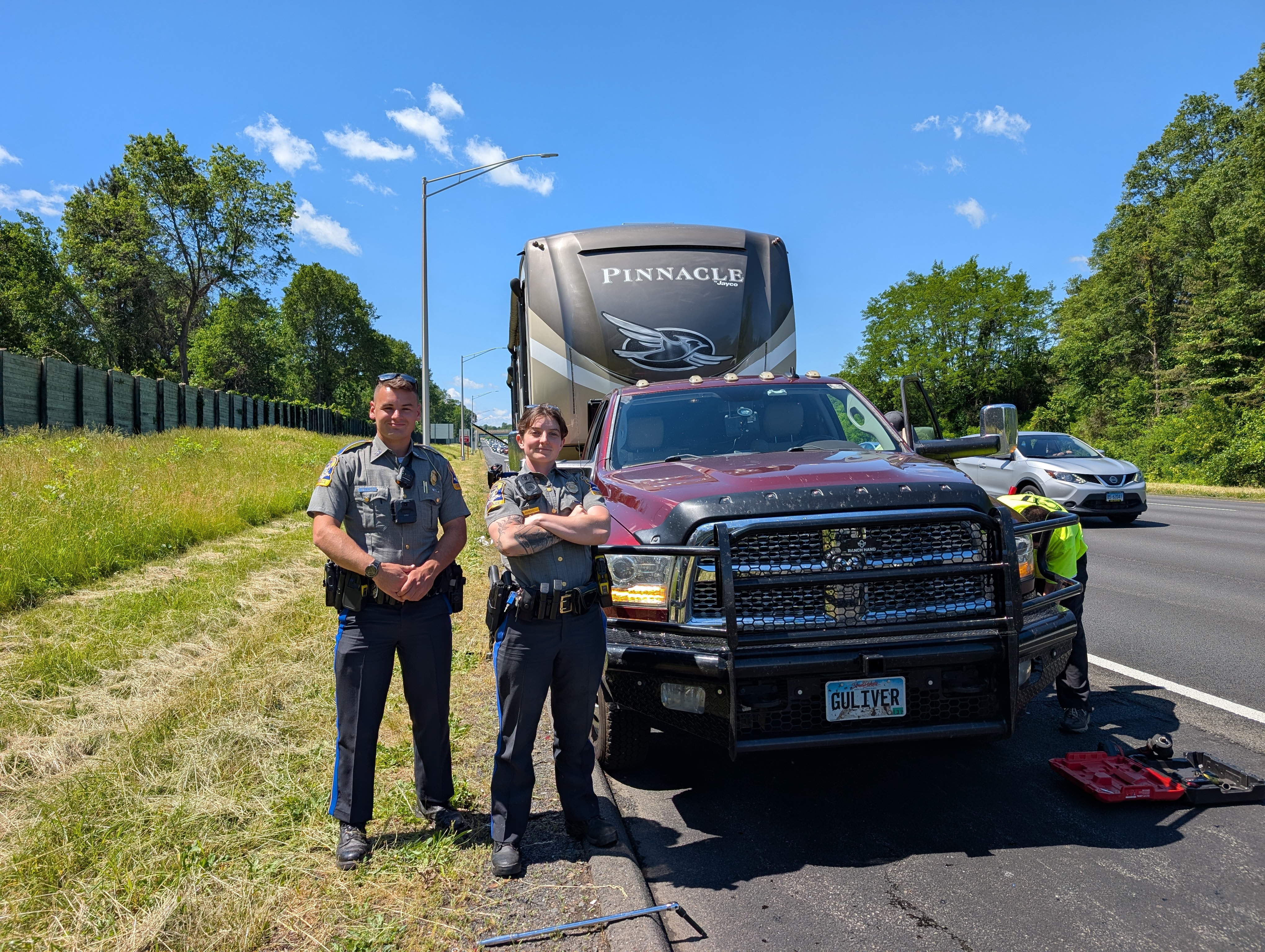 Two Connecticut state troopers next to a truck and fifth wheel on the side of a freeway while a service tech works on a tire
