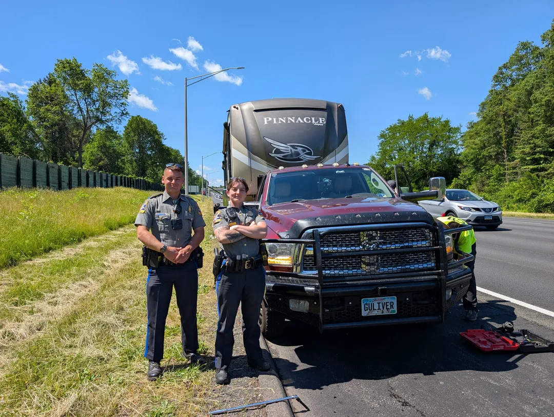 Two Connecticut state troopers next to a truck and fifth wheel on the side of a freeway while a service tech works on a tire