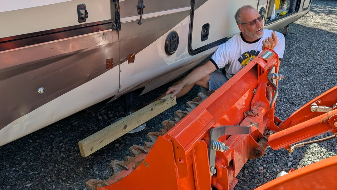 Bob holding a board in front of an orange tractor bucket under a fifth wheel RV
